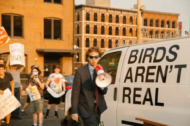 Person speaking through a megaphone beside a Birds Aren't Real sign