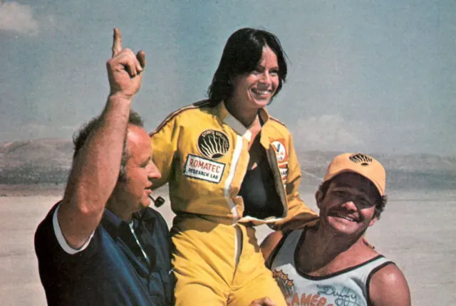 Kitty O'Neil in racing gear, smiling and raising one finger.