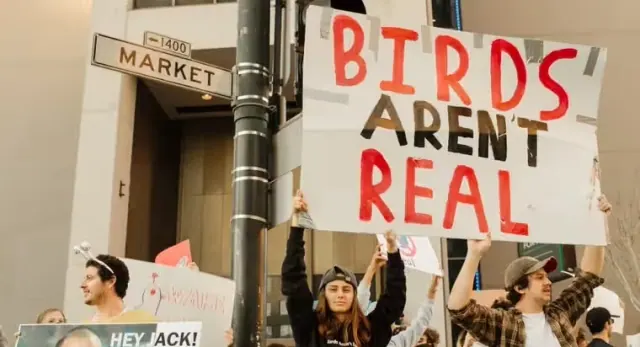 Hand-lettered Birds Aren't Real sign above a street protest