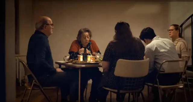 A family sits around a fold out table on fold out chairs in a run down apartment in
