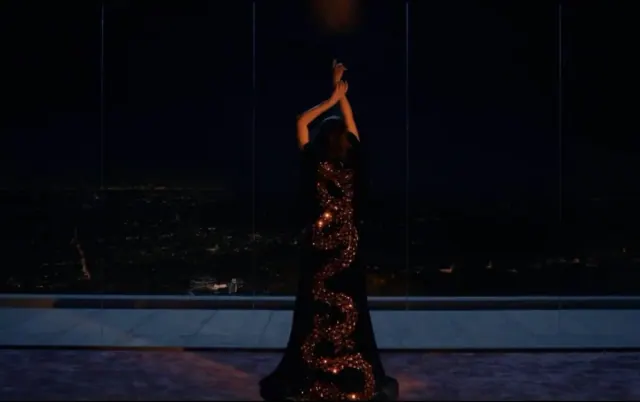Demi Moore in The substance in a long sparkly gown in front of a floor to ceiling window with arms up