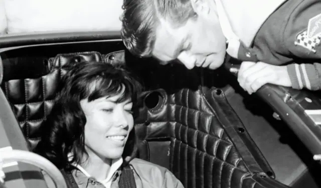 Black-and-white archive photograph of Kitty O'Neil seated in a vehicle with another person nearby.
