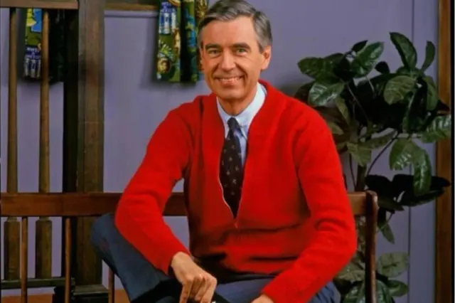 Fred Rogers seated in a red cardigan on the Neighborhood set