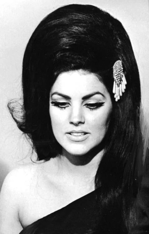 Priscilla looking downward beneath a high dark bouffant